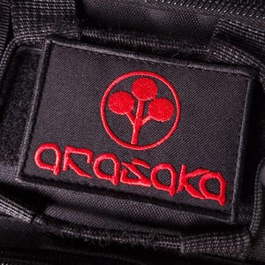 Puede incluir: Un parche rectangular de tela negra con un logotipo circular rojo bordado que muestra un diseño de árbol de tres círculos sobre el texto rojo bordado Arasaka. El parche está cosido sobre un material negro texturizado con cierres de velcro.