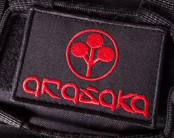 May include: A rectangular black fabric patch featuring an embroidered red circular logo with a three-circle tree design above the red embroidered text Arasaka. The patch is stitched onto a black textured material with visible hook and loop fasteners.