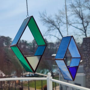 May include: Two hanging stained glass diamond-shaped ornaments. One is green and blue, the other is blue. Both are suspended by chains. The ornaments are hanging in front of a window with a view of trees and a lake.
