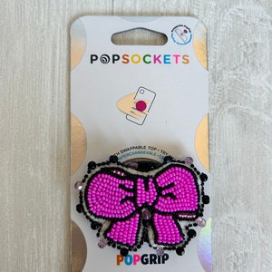 May include: Pink beaded bow PopGrip phone accessory with a black outline and a white background. The packaging includes the text "PopSockets" and "PopGrip".