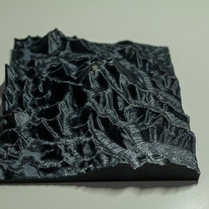May include: A 3D printed model of a mountain range, rendered in a dark grey colour. The model is detailed with ridges and valleys, creating a realistic representation of a mountainous landscape.