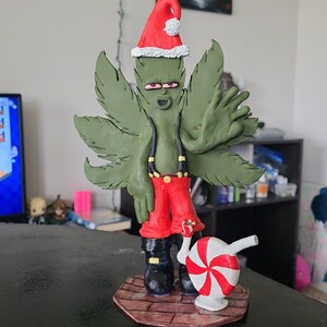 May include: A whimsical sculpture of a cannabis leaf character wearing a Santa hat, red pants, and black boots. The figure has a candy cane-shaped accessory. The sculpture is on a wooden base.