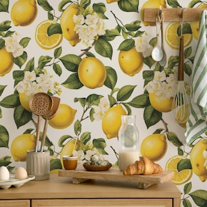 Könnte beinhalten: Eine Küchentapete mit einem Zitronenmotiv. Die Tapete hat einen weißen Hintergrund mit grünen Blättern und gelben Zitronen. Auf der Tapete befinden sich auch weiße Blüten. Die Tapete ist in einer Küchenumgebung mit einer Holzarbeitsplatte, einem Glaskrug, einem Holzschneidebrett und einer Schüssel mit Eiern zu sehen.