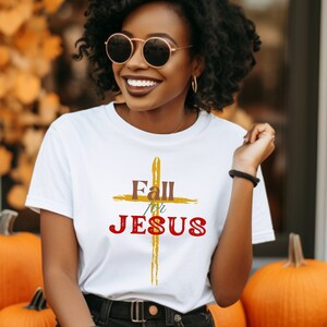 May include: White t-shirt with the text "Fall for Jesus" in red and gold, featuring a cross design. The shirt is worn with black shorts. Pumpkins and fall foliage are in the background.