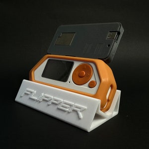 Puede incluir: Un soporte blanco impreso en 3D con la palabra "FLIPPER" impresa en él. Un pequeño dispositivo de mano naranja y blanco está sentado en el soporte. Un dispositivo rectangular negro está sentado en la parte superior del dispositivo naranja y blanco.