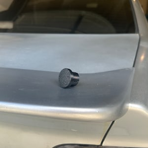 May include: A black plastic push-in fastener with a textured top. The fastener is designed to hold a car part in place.