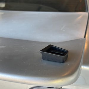 May include: A black plastic car part with a slot in the middle. The part is sitting on a silver car.