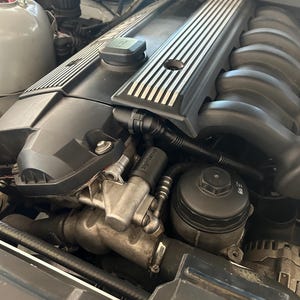 May include: Close-up view of a car engine compartment. The engine is black with silver accents. The engine has a black plastic cover with a silver stripe pattern. There is a black oil filter and various hoses and wires.