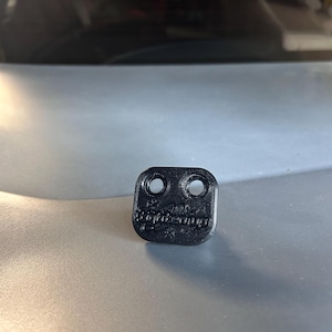 May include: A small, black, rectangular plastic component with rounded corners, featuring two circular holes at the top and the embossed text "tightenup" across the center. The object has a textured surface finish and a flat, low-profile design.