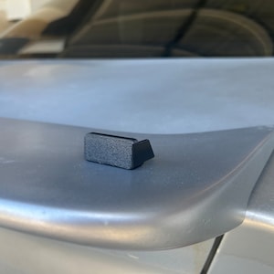 May include: A small, black, rectangular object with a textured surface and a right-angled extension. The object is positioned on a gray surface, likely the trunk of a vehicle. The background is blurred.