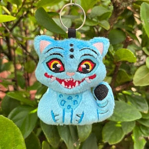 May include: A plush blue cat keychain with embroidered details. The cat has large red eyes with yellow accents, pink ears, and a wide red mouth. It has a black paw and is attached to a metal keyring with a black fabric loop.