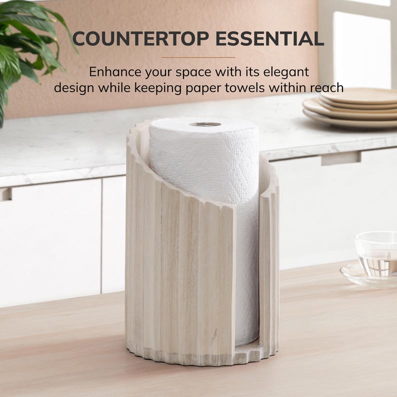 Peut inclure: Un porte-essuie-tout cylindrique en bois clair avec une texture nervur&eacute;e verticale et une large ouverture frontale. Le support contient un rouleau d'essuie-tout blanc. Le texte au-dessus indique : COUNTERTOP ESSENTIAL. Enhance your space with its elegant design while keeping paper towels within reach.