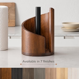 May include: A paper towel holder featuring a curved, dark brown wooden base and a central matte black metal rod. The design includes a vertical wooden panel that wraps around the side. The text "Available in 7 finishes" appears below.