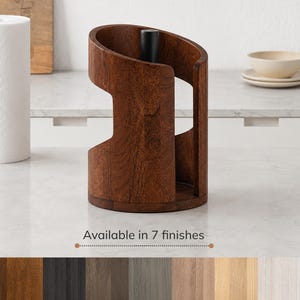 Peut inclure: Un porte-essuie-tout en bois brun foncé de forme cylindrique avec une tige centrale en métal noir. Le support présente une découpe incurvée sur le devant. Sous le support, le texte indique : Available in 7 finishes, avec sept échantillons de bois en rangée.