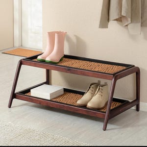 May include: A two-tier shoe rack constructed from dark brown wood with angled legs. Each tier features a black frame containing a woven tan fiber mat. The top shelf holds a pair of light pink rubber boots, while the bottom shelf holds a pair of beige lace-up ankle boots and a small white rectangular box.