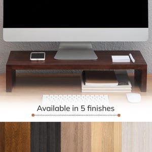 Peut inclure: Un support d'écran en bois brun foncé avec un plateau rectangulaire et deux supports latéraux. Le support accueille un écran d'ordinateur, un smartphone, un bloc-notes et un stylo. Sous le texte Available in 5 finishes figurent cinq échantillons.