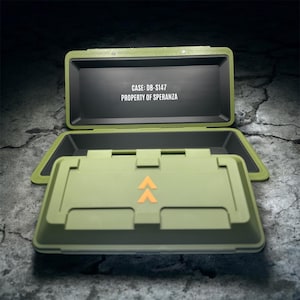 May include: An open, olive-green case with a black interior. The case lid is open, revealing the text "CASE: DB-S147 PROPERTY OF SPERANZA" in white. The bottom section has orange arrow symbols. The case is on a textured, gray surface.