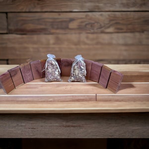 May include: Aromatic cedar wood blocks arranged in an arch, framing two small clear bags filled with dried herbs. The cedar blocks are a rich reddish-brown color, and the bags are tied with white string. The background is a wooden surface.
