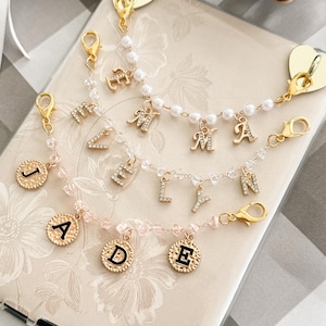 May include: Three gold-toned chain charms featuring lobster clasps. One chain has white faux pearls and rhinestone letters spelling EMMA. Another has clear beads and rhinestone letters spelling ELYN. The third has pink beads and gold coin charms with black letters spelling JADE.