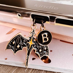 May include: A black and gold charm set featuring a bat and a circular pendant with the letter "B". The bat charm has intricate wing details and a yellow star. The charms are attached to a black cord.