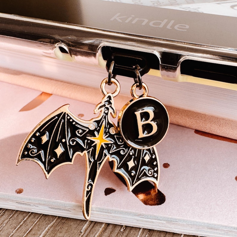 May include: A black and gold charm set featuring a bat and a circular pendant with the letter "B". The bat charm has intricate wing details and a yellow star. The charms are attached to a black cord.