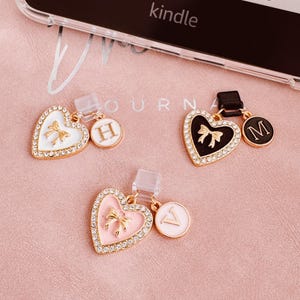 Könnte beinhalten: Drei herzförmige Handy-Anhänger mit goldfarbenen Verzierungen und Strass-Akzenten. Einer ist weiß mit einer goldenen Schleife, einer ist schwarz mit einer goldenen Schleife und einer ist rosa mit einer goldenen Schleife. Jeder Anhänger hat einen kleinen, runden Initialen-Anhänger. Das Wort "kindle" ist sichtbar.