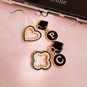 May include: Gold-toned phone charms with black accents. One charm is a heart shape, another is a four-leaf clover, and two are round with the letters "P" and "C". Each charm is embellished with clear crystals.