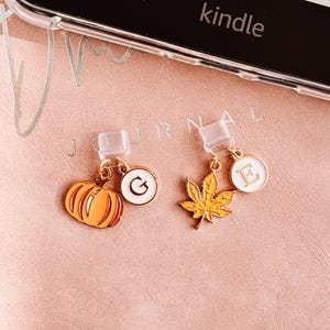 May include: Two phone charms with gold-colored hardware. One charm is an orange pumpkin, and the other is a yellow maple leaf. Both charms have white circular pendants with gold letters. The word "kindle" is visible.