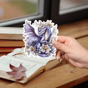 May include: A purple dragon with gold accents is surrounded by white flowers. The dragon has its wings spread out and is looking to the left. The sticker is on a white background.