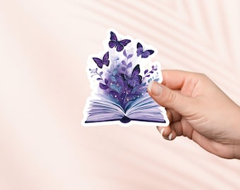 Puede incluir: Una pegatina que representa un libro abierto con páginas moradas y un lomo oscuro. Mariposas y follaje en tonos morados y azules emergen del libro. La pegatina está sujeta por una mano.