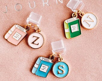 Op de afbeelding: Drie kleurrijke, boekvormige bedels met goudkleurige accenten en initiaalhangers. De bedels zijn roze, groen en blauw, elk gecombineerd met een ronde hanger met een letter. Het woord "JOURNAL" is zichtbaar op de achtergrond.