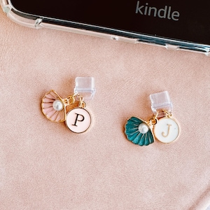 Könnte beinhalten: Zwei Handy-Anhänger mit goldfarbenen Akzenten. Ein Anhänger zeigt eine rosa Muschel mit einer Perle und einen weißen Kreis mit dem Buchstaben "P". Der andere Anhänger hat eine türkisfarbene Muschel mit einer Perle und einen weißen Kreis mit dem Buchstaben "J".