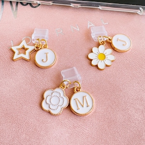 Puede incluir: Tres colgantes dorados para teléfono con detalles de esmalte blanco. Un colgante presenta una estrella, otro una margarita y el tercero una forma de flor. Cada colgante tiene una letra inicial a juego y un clip de plástico transparente.
