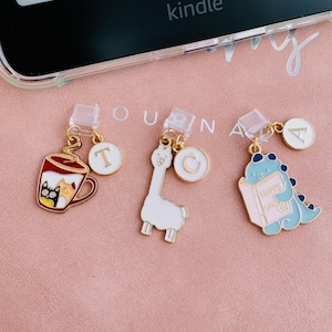 May include: Three decorative phone charms with gold-colored accents. One charm is a red coffee cup with a black cat, another is a white llama, and the last is a blue dinosaur reading a book with the word "Happy". Each charm has a small white circle with a letter.