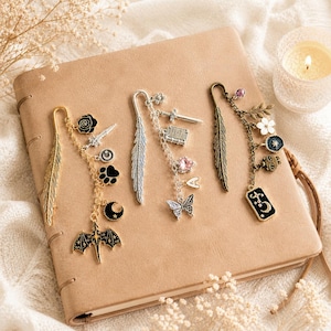 May include: A collection of decorative metal bookmarks with dangling charms, including feathers, swords, butterflies, and celestial motifs. The bookmarks are gold, silver, and bronze, resting on a tan leather-bound journal.