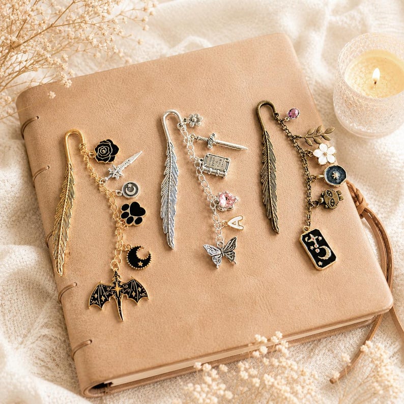 May include: A collection of decorative metal bookmarks with dangling charms, including feathers, swords, butterflies, and celestial motifs. The bookmarks are gold, silver, and bronze, resting on a tan leather-bound journal.