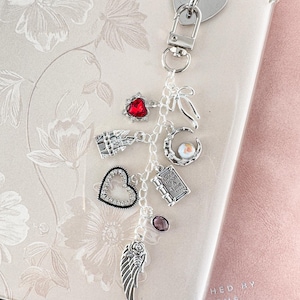 Op de afbeelding: Een zilveren bedel sleutelhanger met meerdere decoratieve bedels. Bedels zijn onder meer een rood hart, een helder hart, een vleugel, een boek en een maan. De sleutelhanger is bevestigd aan een doorzichtige telefoonhoes.