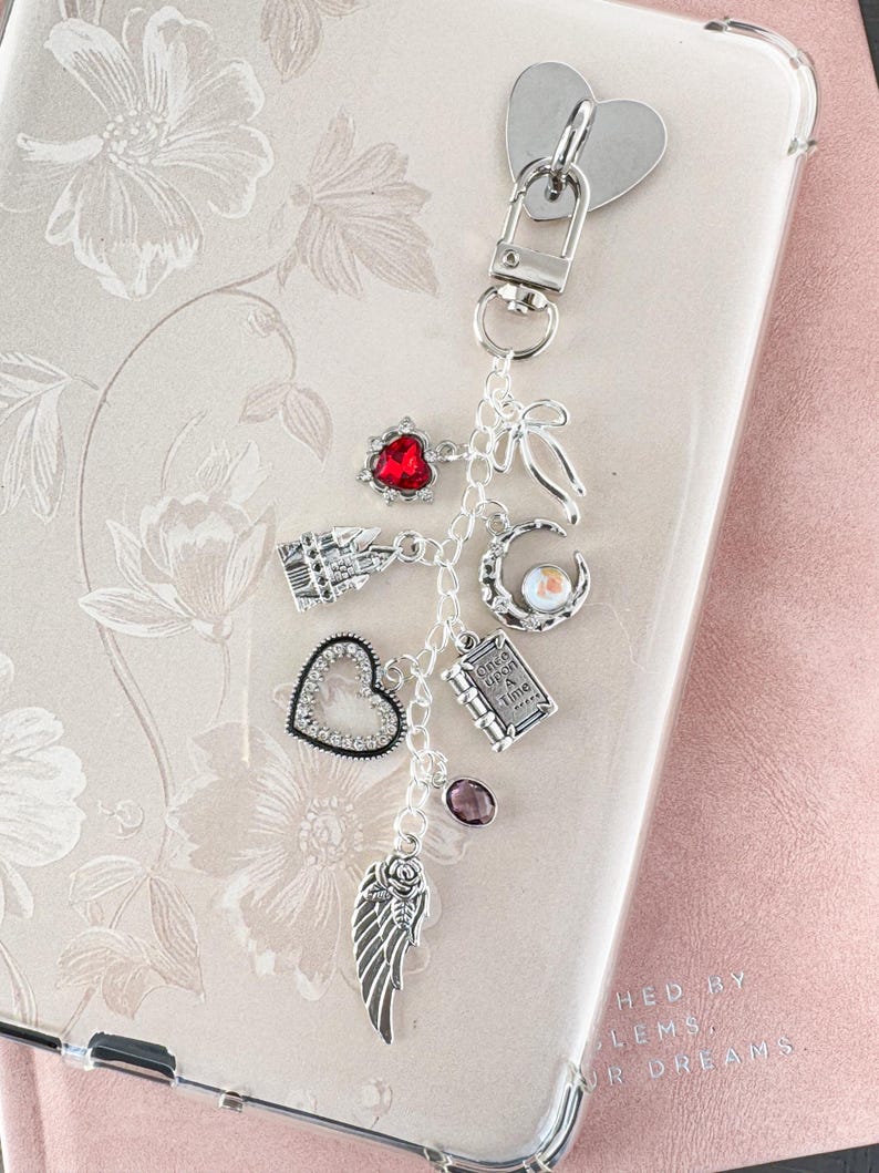 Op de afbeelding: Een zilveren bedel sleutelhanger met meerdere decoratieve bedels. Bedels zijn onder meer een rood hart, een helder hart, een vleugel, een boek en een maan. De sleutelhanger is bevestigd aan een doorzichtige telefoonhoes.