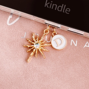 Op de afbeelding: Goudkleurige zonnestraalbedel met een centrale iriserende edelsteen en een witte emaille bedel met de letter "D" die aan een doorzichtige telefoonhoes hangt. Het woord "kindle" is zichtbaar op de telefoon.