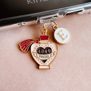 May include: A gold-colored charm with a pink heart-shaped bottle design, labeled "LOVE POTION" with a banner. A white circular charm with a gold letter "E" is also attached. The charm is hanging from a clear phone case.