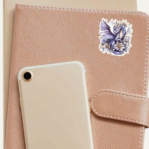 May include: A sticker featuring a purple dragon with wings spread, surrounded by white flowers. The dragon is perched on a branch with its head turned to the side.