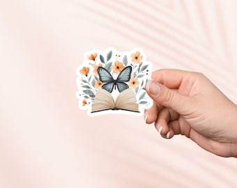 Puede incluir: Una pegatina con un libro abierto con una mariposa azul posada encima. El libro está rodeado de delicadas flores de acuarela en tonos naranja y amarillo.