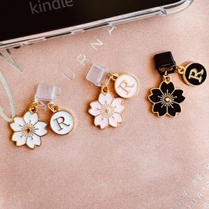 Könnte beinhalten: Drei Handy-Anhänger mit floralen und Initialen-Designs. Ein Anhänger zeigt eine schwarze Blume, ein weiterer eine rosa Blume und der dritte eine weiße Blume. Jeder Blumenanhänger hat einen goldfarbenen Rand und ist mit einem runden Anhänger mit dem Buchstaben "R" versehen.