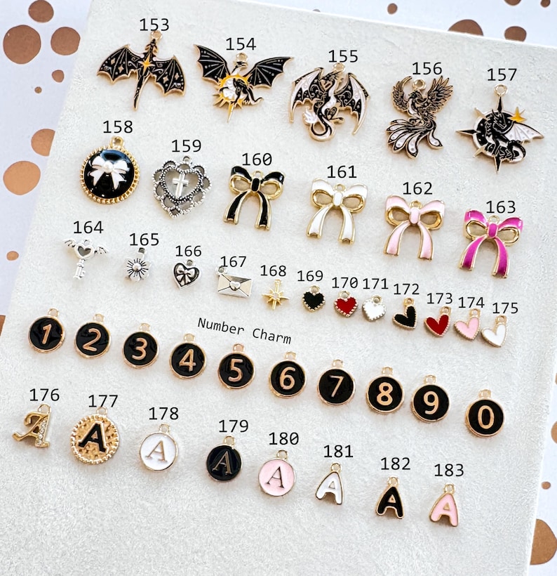 Op de afbeelding: Een verzameling decoratieve bedels met verschillende ontwerpen, waaronder draken, strikken, harten en cijfers. De bedels zijn goud, zwart, wit en roze. Sommige bedels hebben de letter "A".