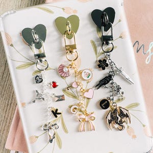 Op de afbeelding: Drie decoratieve sleutelhangers met hartvormige sluitingen. E&eacute;n sleutelhanger heeft een strik, een hart en een drakenbedel. Een andere heeft een sleutel, een schelp en een hartbedel. De derde heeft een klavertje vier, een zwaard en een ravenbedel. Elke sleutelhanger heeft een sluiting in een andere kleur.