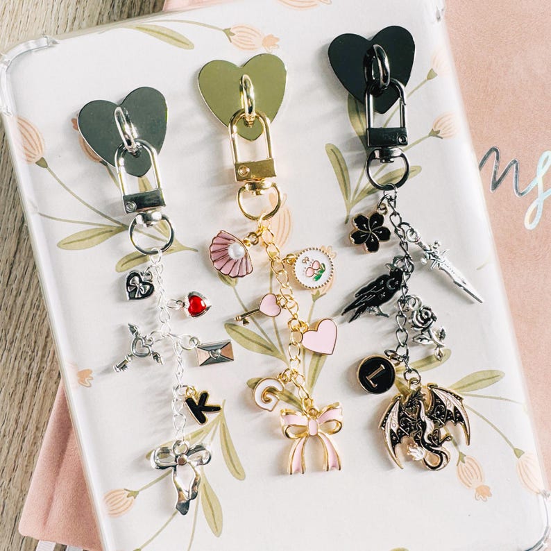 Op de afbeelding: Drie decoratieve sleutelhangers met hartvormige sluitingen. E&eacute;n sleutelhanger heeft een strik, een hart en een drakenbedel. Een andere heeft een sleutel, een schelp en een hartbedel. De derde heeft een klavertje vier, een zwaard en een ravenbedel. Elke sleutelhanger heeft een sluiting in een andere kleur.
