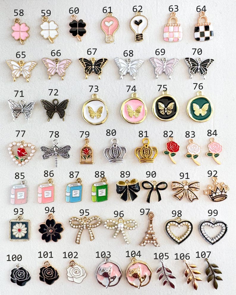 Op de afbeelding: Een verzameling kleine, decoratieve bedels in verschillende vormen en ontwerpen. Inclusief vlinders, bloemen, kronen, strikken, boeken, harten en Eiffeltoren bedels. De bedels zijn in goud, zilver, zwart, roze en wit.