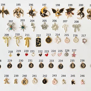 May include: A collection of small, decorative charms in various shapes and designs. Includes charms shaped like dragons, bows, flowers, hearts, and letters. The charms are in gold, black, white, and pink colors.
