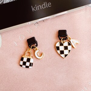 May include: Two decorative charms with black and white checkered patterns. One charm is a handbag, the other is a heart. Both charms have gold accents and hang from a black clasp. The word "kindle" is visible in the background.