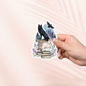May include: A sticker depicting a black wolf and a white wolf on a pile of books, with a raven in flight. The design incorporates a starry night, pine trees, and lavender. The sticker is held by a hand.
